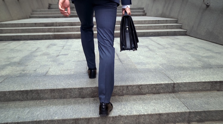 View of a grey staircase where legs in a blue suit and an arm holding a black briefcase walk up the steps.