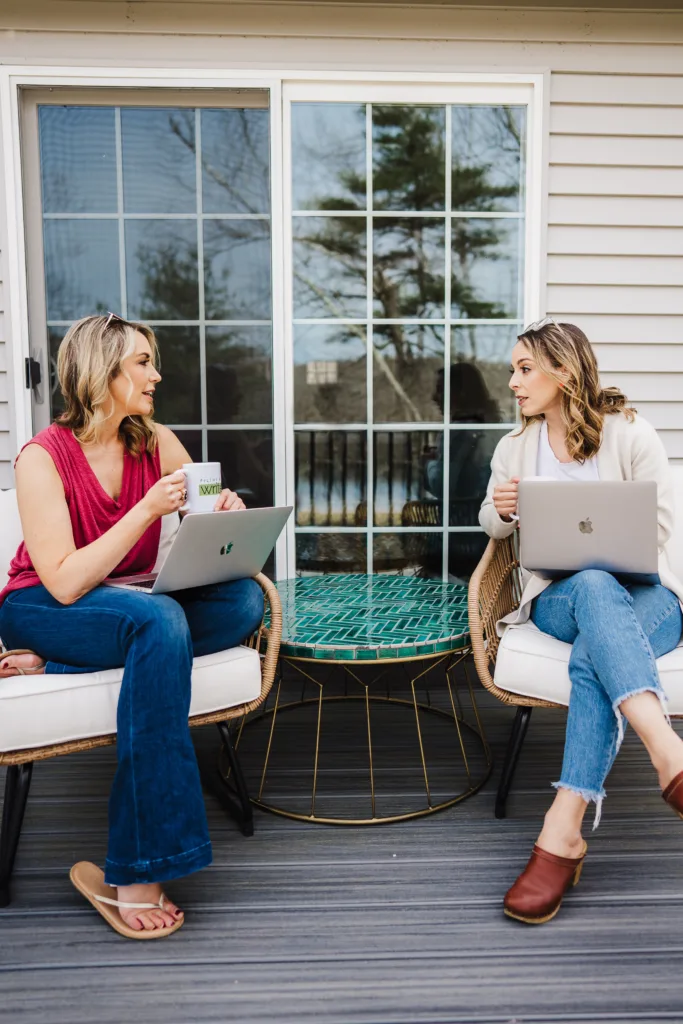 Nicki and Kate, hosts of the Build Your Copywriting Business podcast, sit outside with laptops on their laps and holding Filthy Rich Writer mugs in their hands.