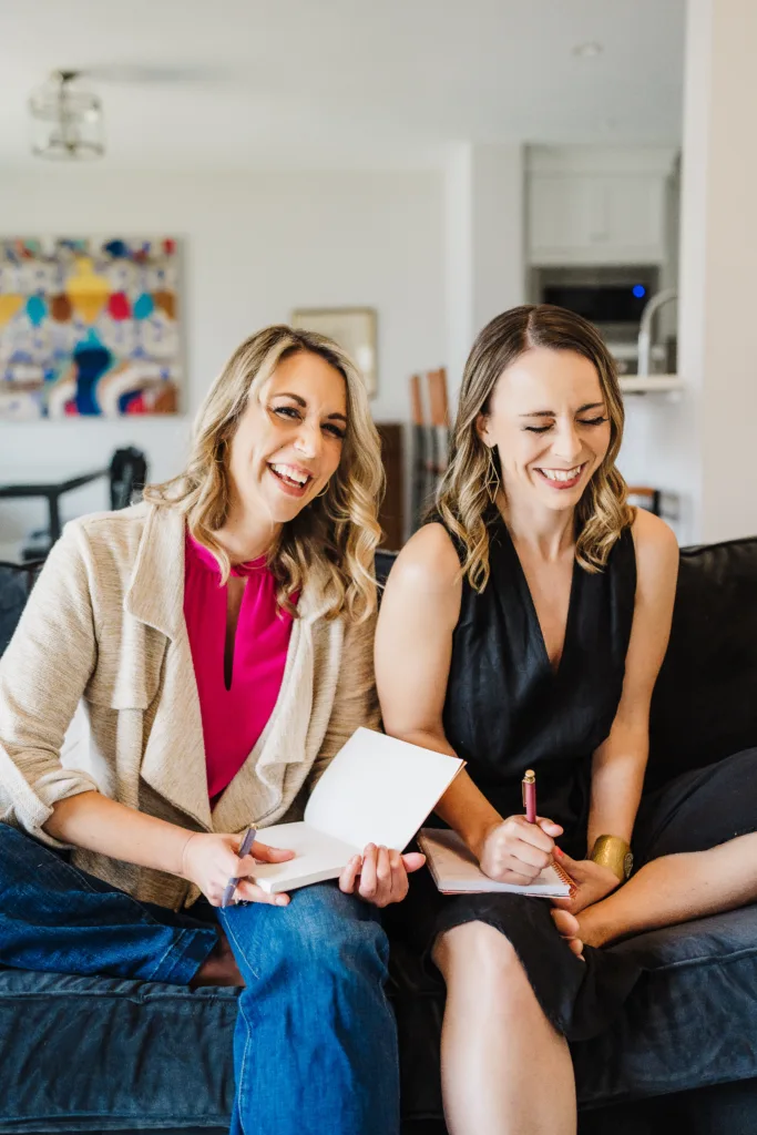 Nicki and Kate, hosts of the Build Your Copywriting Business podcast, sit next to each other on a couch laughing with notebooks in their laps.