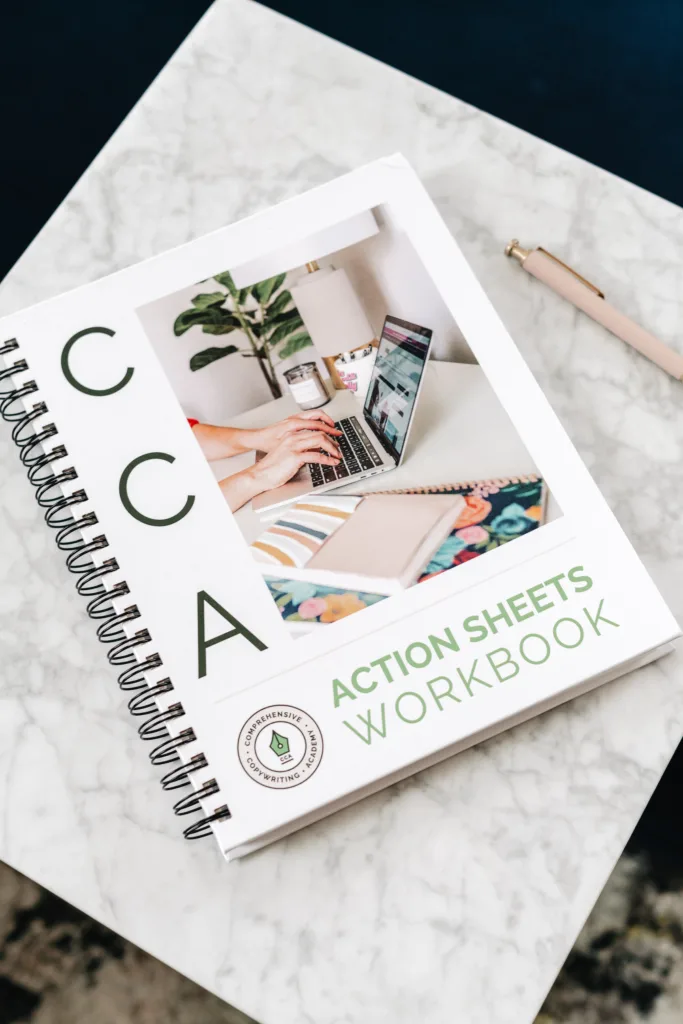 A spiral-bound workbook that says CCA Action Sheets Workbook sits on a table beside a pen.