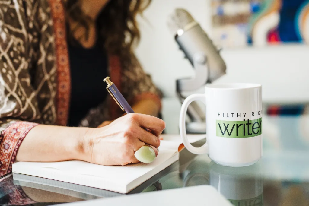 Filthy Rich Writer mug sits on desk while Nicki's hand writes in notebook.