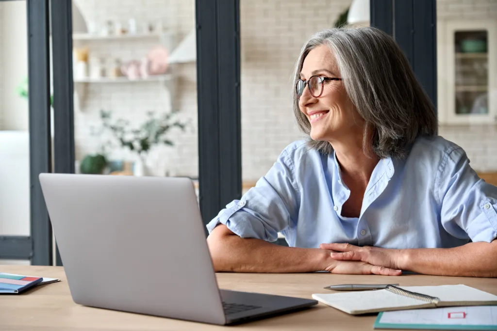 Woman with grey hair and glasses smiles while glancing away from her laptop that is open in front of her on the desk.
