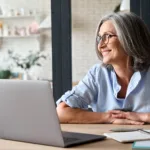 Woman with grey hair and glasses smiles while glancing away from her laptop that is open in front of her on the desk.