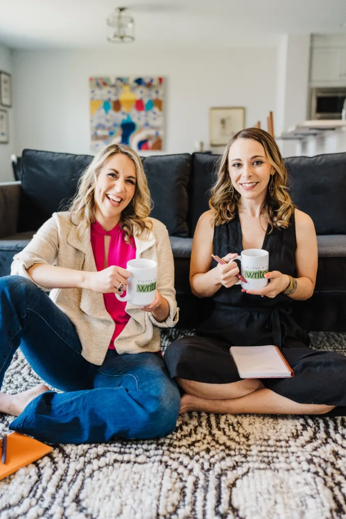Nicki Krawczyk and Kate Sitarz, hosts of the Build Your Copywriting Business podcast, hold mugs that say "Filthy Rich Writer" as they sit side-by-side in front of a couch