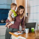 Woman stands, holding a small child in one arm while typing on laptop at the kitchen table with the other.