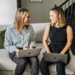 Nicki Krawczyk, founder of Filthy Rich Writer, and Kate Sitarz, head copywriting coach in the Comprehensive Copywriting Academy, sit on a couch with laptops on their laps.