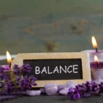 Sign with the word "balance" on in with purple flowers and lit purple candles surrounding it