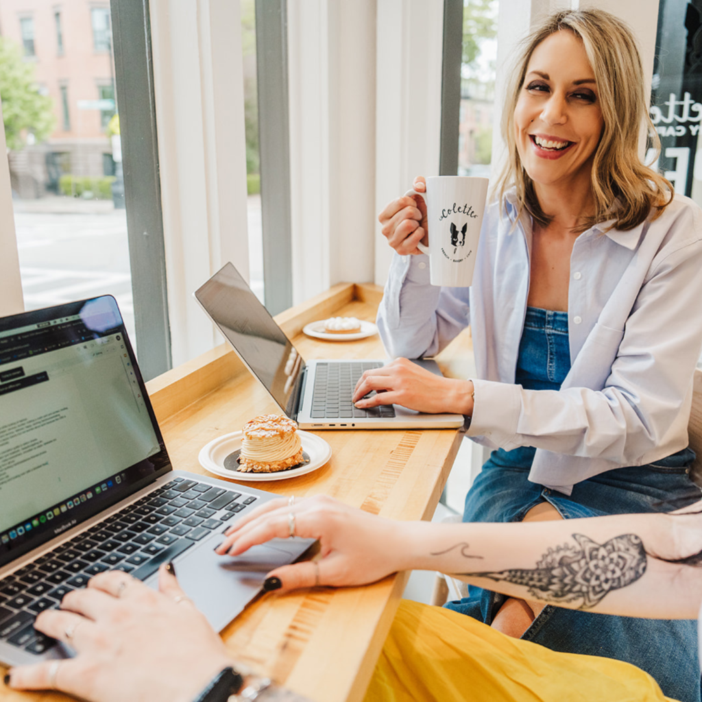 Nicki Krawczyk, founder of Filthy Rich Writer and the Comprehensive Copywriting Academy, holds a cup of coffee while someone types on a laptop in the foreground.