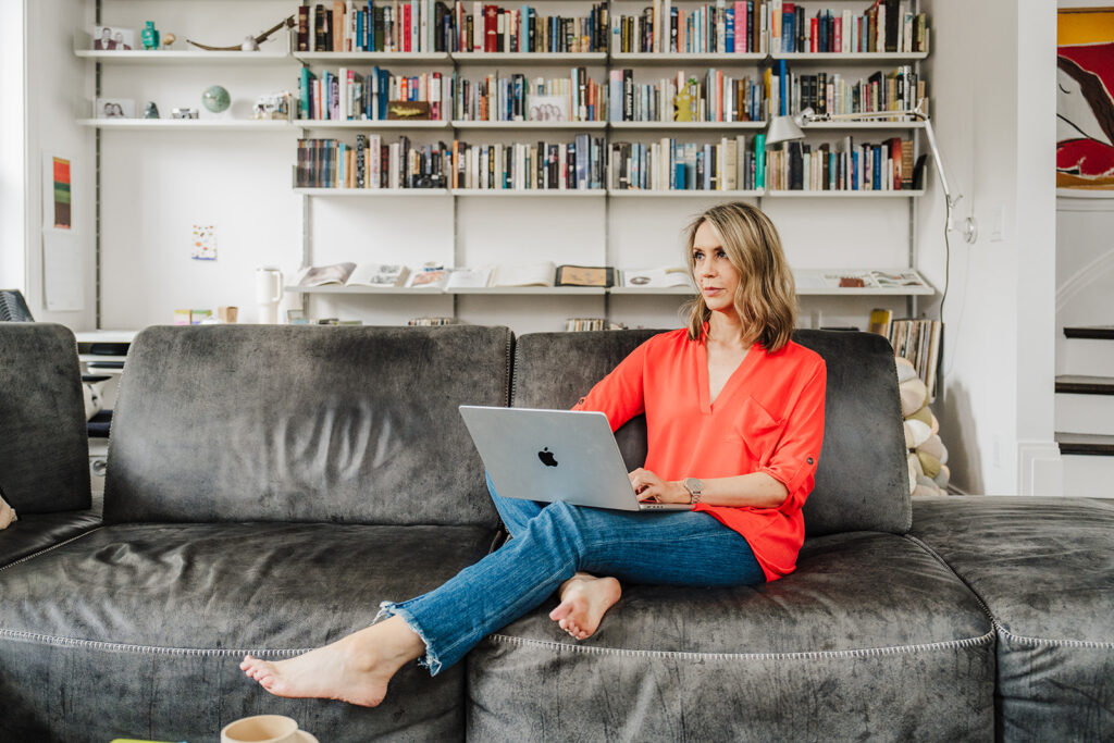 Nicki sits on a black leather couch wearing a bright orange top and jeans with a laptop on her lap and a bookshelf in the backdrop.