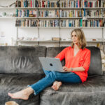 Nicki sits on a black leather couch wearing a bright orange top and jeans with a laptop on her lap and a bookshelf in the backdrop.