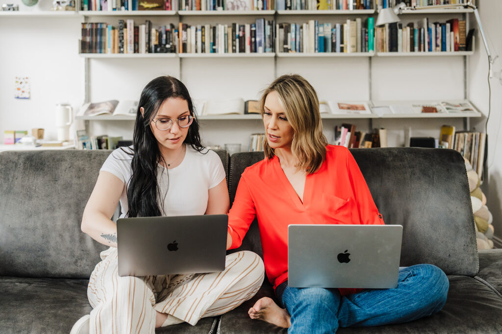 Nicki Krawczyk, founder of the Comprehensive Copywriting Academy, points over the shoulder of a collaborator, both with laptops on their laps while sitting on the couch.