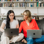 Nicki Krawczyk, founder of the Comprehensive Copywriting Academy, points over the shoulder of a collaborator, both with laptops on their laps while sitting on the couch.