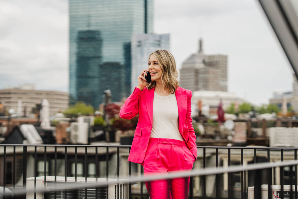 Photo of Nicki, founder of Filthy Rich Writer and the Comprehensive Copywriting Academy, in a hot pink blazer and pants talking on the phone with the Boston skyline in the background.