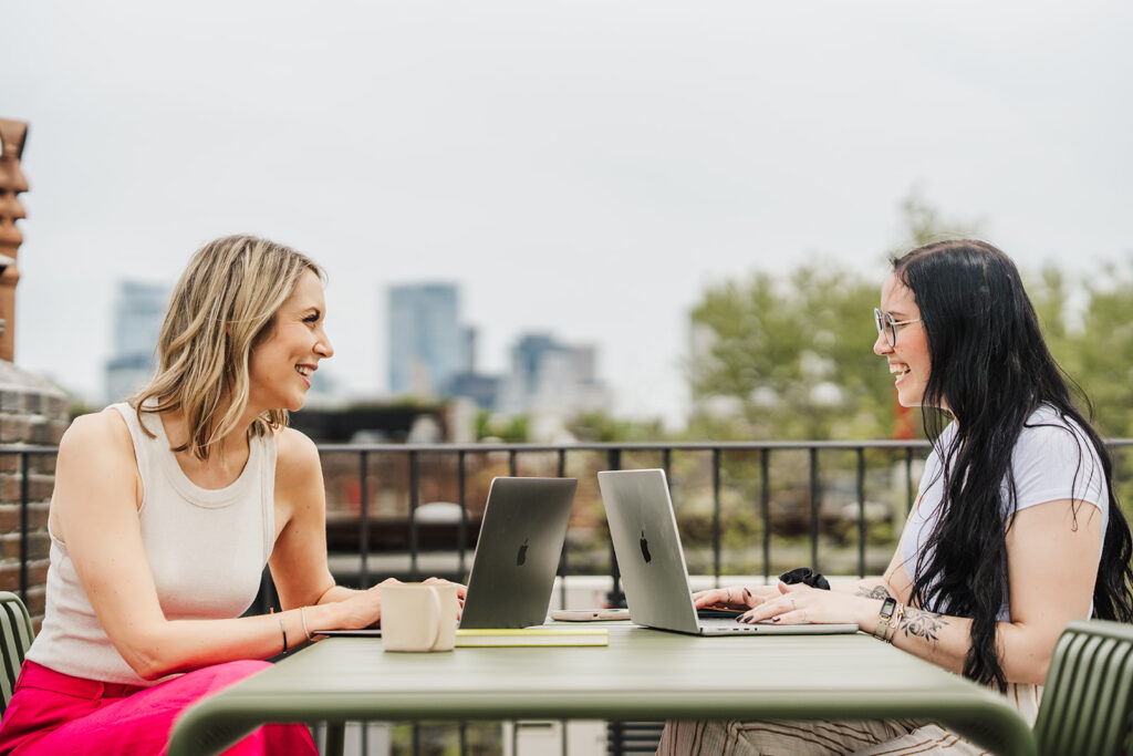 Nicki Krawczyk, founder of Filthy Rich Writer and the Comprehensive Copywriting Academy, chats with designer Meaghan with their laptops open on a roof deck overlooking the Boston skyline.