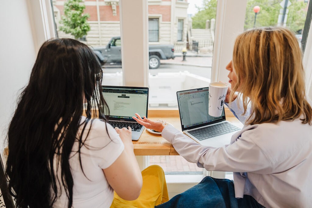 Nicki Krawczyk, founder of Filthy Rich Writer and the Comprehensive Copywriting Academy, has a laptop in front of her at a coffeeshop and gestures to designer Meaghan's screen with one hand while holding a cup of coffee with the other.