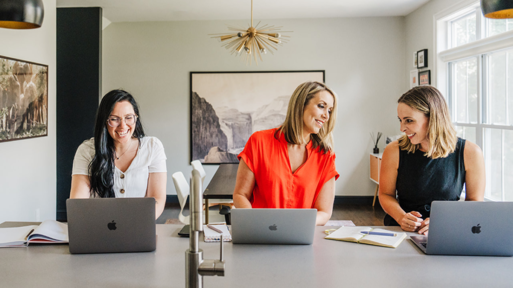 Nicki Krawczyk, founder of the Comprehensive Copywriting Academy works with team members, laptops open, at a kitchen table.