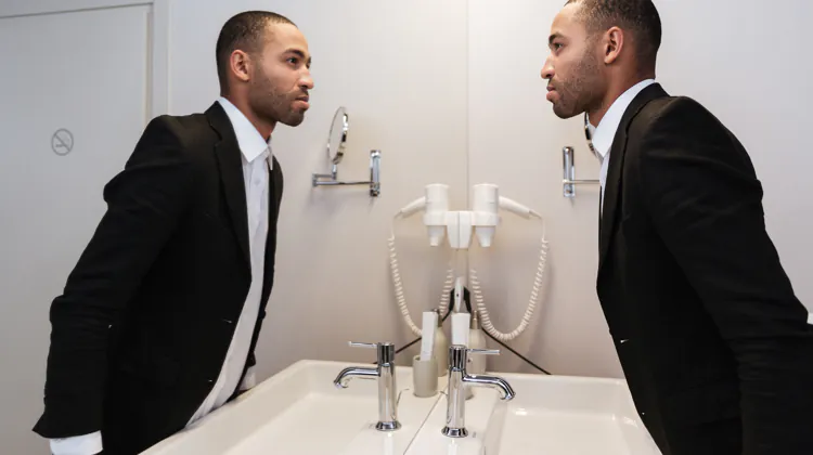 Black man with close-cut haircut stand sin a tux in front of a sink looking into a mirror with his reflection looking back.