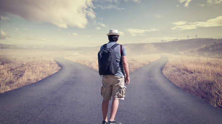 A man wearing a white fedora, blue shirt, and khaki shorts stands at a fork in the road in the middle of prairie land. He is carrying a navy backpack.