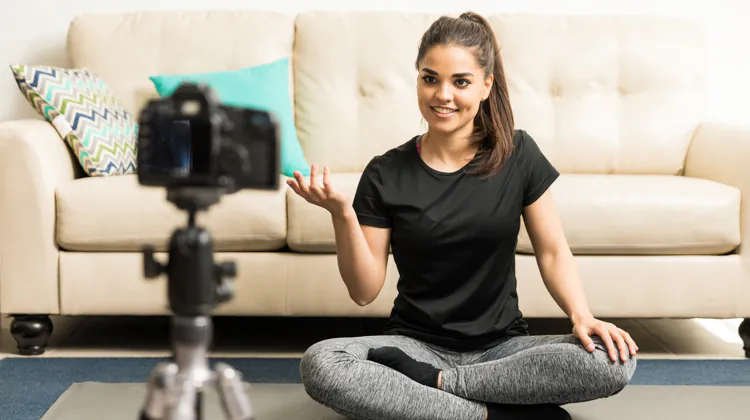 A woman is sitting cross-legged on the floor in front of a cream colored couch. She is looking at a camera mounted on a tripod. The woman is wearing a black shirt and gray leggings.