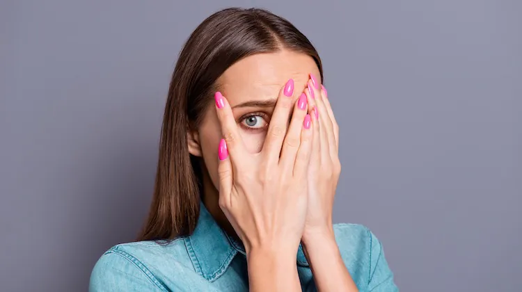 Woman in denim top and Barbie-pink nails covers her face with her hands with one eyeball peeking through one hand.